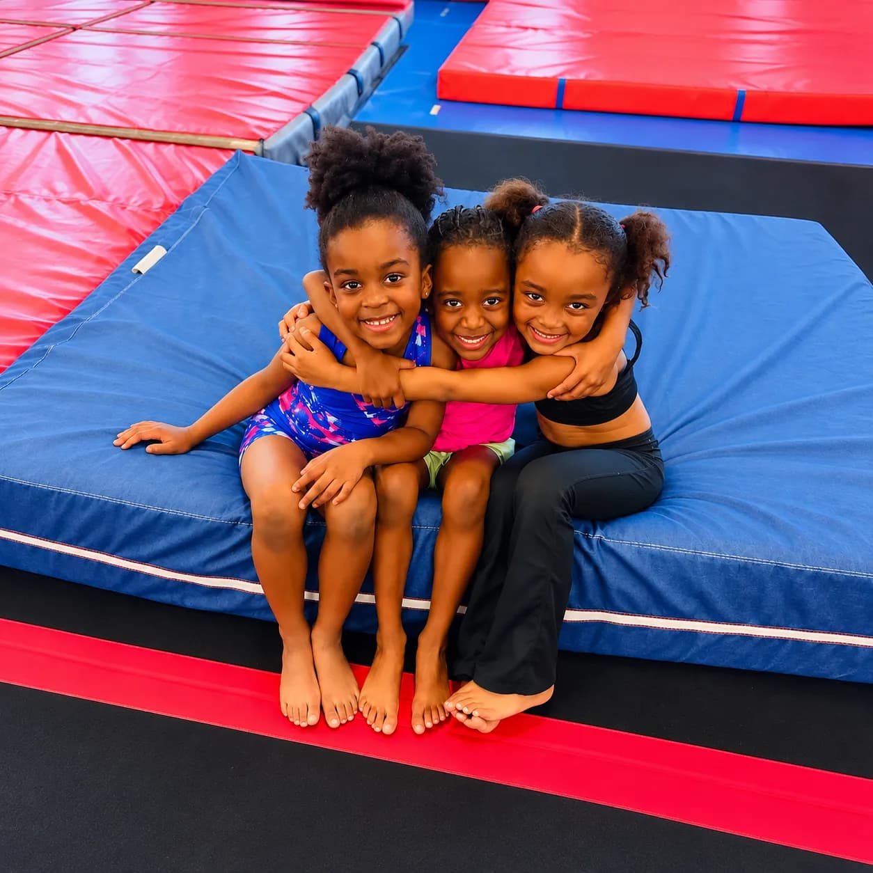 Three young girls hugging and smiling while sitting on a blue mat