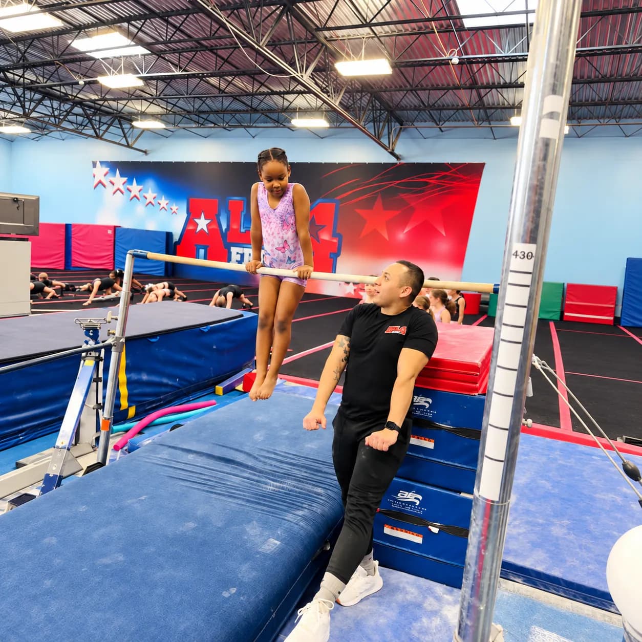 A young gymnast holding herself up on a low bar as a coach stands beside her
